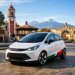 Fotografía realista de un prototipo de auto eléctrico compacto con colores de la bandera de México circulando por una plaza en Tlaxcala, con montanas al fondo